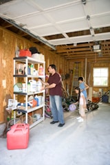 A garage with wooden walls and an open door is partially filled with various items. A man stands near metal shelves, organizing or picking up cleaning products and other household supplies. Behind him, two children stand, observing or assisting. The floor is cluttered with toys like a basketball, a scooter, and some gardening tools.