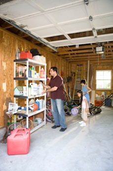 A garage with wooden walls and an open door is partially filled with various items. A man stands near metal shelves, organizing or picking up cleaning products and other household supplies. Behind him, two children stand, observing or assisting. The floor is cluttered with toys like a basketball, a scooter, and some gardening tools.