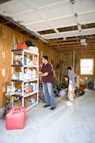 A garage with wooden walls and an open door is partially filled with various items. A man stands near metal shelves, organizing or picking up cleaning products and other household supplies. Behind him, two children stand, observing or assisting. The floor is cluttered with toys like a basketball, a scooter, and some gardening tools.