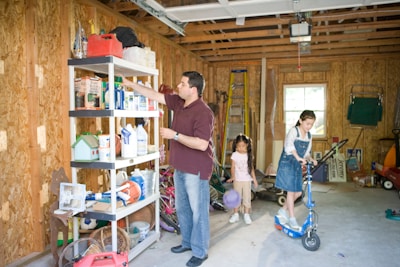 A man is organizing items on a shelf inside a garage. Two children, one holding a ball and the other on a scooter, are nearby. The garage contains various items such as bottles, tools, and sports equipment. The walls are unfinished wood, and there's a small window allowing natural light in.