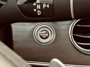 A close-up of a car interior shows a wooden dashboard panel, featuring a circular engine start/stop button. The steering wheel is partially visible, and a gear shift indicator displays the letters P, R, N, D.