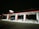 A gas station at night with bright overhead lights illuminating several fuel pumps. The ground appears wet or icy, reflecting the lights above, and there is a van parked by one of the pumps. The overall setting is quiet with no people visible, and the surroundings are dark.