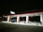 A gas station at night with bright overhead lights illuminating several fuel pumps. The ground appears wet or icy, reflecting the lights above, and there is a van parked by one of the pumps. The overall setting is quiet with no people visible, and the surroundings are dark.