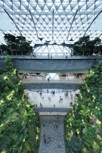 A modern indoor space featuring multiple levels with a glass roof composed of geometric patterns. Greenery lines the walls, creating a lush vertical garden. People are walking through the open atrium, giving the environment a lively and bustling atmosphere.