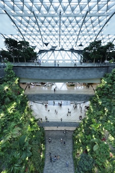 A modern indoor space featuring multiple levels with a glass roof composed of geometric patterns. Greenery lines the walls, creating a lush vertical garden. People are walking through the open atrium, giving the environment a lively and bustling atmosphere.