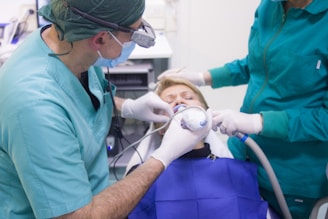 A dental procedure in progress with two medical professionals attending to a patient. The patient is lying back in a dental chair, wearing a blue protective bib. One dentist is using a dental instrument while the other assists, both wearing surgical masks and gloves.