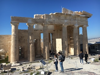 A group of people stands in front of a historic ancient Greek temple with tall, weathered columns under a clear blue sky. The structure appears to be partially restored, with stone debris and informational signs nearby.