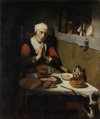 An elderly woman with her hands clasped in prayer sits at a table featuring a variety of food items, including bread, butter, and a pitcher. The setting is dimly lit, with a cat stretching toward the table.