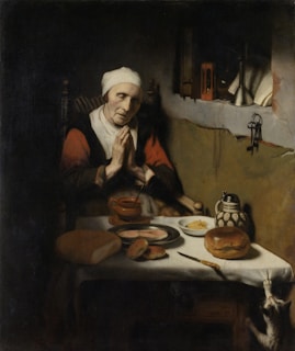 An elderly woman with her hands clasped in prayer sits at a table featuring a variety of food items, including bread, butter, and a pitcher. The setting is dimly lit, with a cat stretching toward the table.