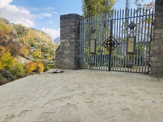 A gated entrance stands at the forefront, made of vertical metal bars with decorative patterns and flanked by stone pillars. Beyond the gate lies a scenic view of a hilly landscape with colorful autumn foliage, showcasing a mix of green, yellow, and orange trees. The background features distant mountains under a partly cloudy sky.