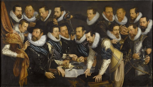 A group of men dressed in elaborate, historical clothing gather around a table with silverware and dishes. The men have ruffled collars and varying expressions, standing in a dimly lit setting that highlights their fine attire and facial details. Each individual seems engaged in conversation or action, holding cups, plates, or gesturing with hands.