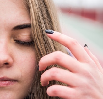 A close-up view of a person's face with their eyes closed and a hand touching their hair. The person has long hair and appears to be in a state of calm or introspection. The background is blurred, emphasizing the focus on the face and hand.