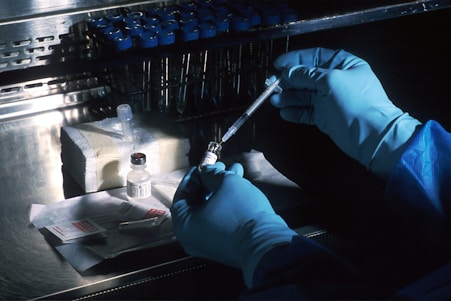 A person wearing blue protective gloves is handling a syringe and a vial on a metallic surface. The background includes a stainless steel shelf holding rows of small bottles with blue caps. There is also a pack of alcohol wipes and a box containing laboratory equipment.
