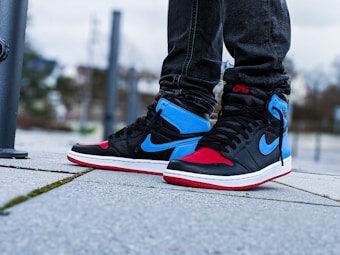 Sneakers with a prominent blue and red color scheme, featuring a distinctive logo on the side. The shoes are worn with dark jeans, and the scene is set on a concrete pavement with a blurred urban background.