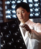 A person in a white lab coat is examining X-ray images using a pen, with illuminated X-ray scans visible in the background. The setting suggests a medical or diagnostic environment, possibly a hospital or clinic.