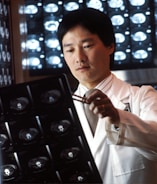 A person in a white lab coat is examining X-ray images using a pen, with illuminated X-ray scans visible in the background. The setting suggests a medical or diagnostic environment, possibly a hospital or clinic.