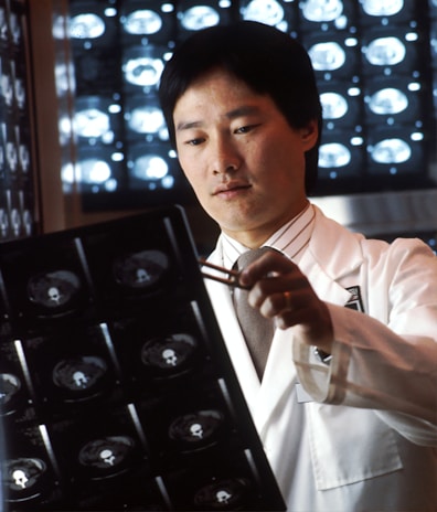 A person in a white lab coat is examining X-ray images using a pen, with illuminated X-ray scans visible in the background. The setting suggests a medical or diagnostic environment, possibly a hospital or clinic.