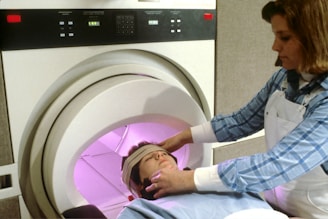 A person is undergoing a medical scan inside an MRI machine, with another individual assisting by adjusting their position. The scene appears clinical, with the MRI machine's interior emitting a soft pink glow. The assistant is wearing a blue checkered shirt and a white apron.