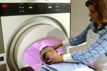 A person is undergoing a medical scan inside an MRI machine, with another individual assisting by adjusting their position. The scene appears clinical, with the MRI machine's interior emitting a soft pink glow. The assistant is wearing a blue checkered shirt and a white apron.
