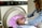 A person is undergoing a medical scan inside an MRI machine, with another individual assisting by adjusting their position. The scene appears clinical, with the MRI machine's interior emitting a soft pink glow. The assistant is wearing a blue checkered shirt and a white apron.