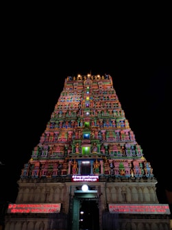 A tall, ornate temple tower richly decorated with colorful sculptures and intricate carvings stands against a dark night sky. The bright illumination highlights various deities and traditional motifs, adding vibrancy to each layer of the structure. Signage with illuminated text in another language is visible near the entrance.