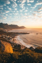 A scenic coastal view featuring a large bay with calm waters and several small waves, a sandy beach lined with buildings and vegetation, and a backdrop of tall, rugged mountains under a partly cloudy sky during sunset. The warm sunlight casts a golden hue over the landscape.