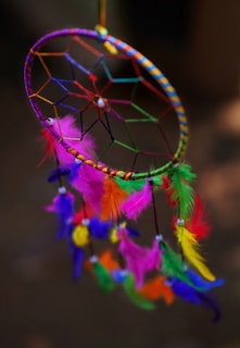 A vibrant dreamcatcher with a woven circular pattern and colorful feathers. The feathers include shades of pink, green, yellow, orange, and purple, hanging from various points on the woven circle. The dreamcatcher is set against a blurred dark background, enhancing the vividness of its colors.