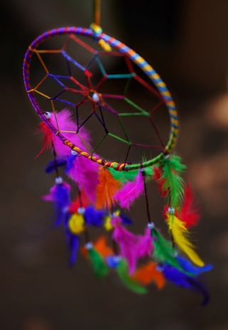 A vibrant dreamcatcher with a woven circular pattern and colorful feathers. The feathers include shades of pink, green, yellow, orange, and purple, hanging from various points on the woven circle. The dreamcatcher is set against a blurred dark background, enhancing the vividness of its colors.