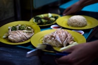 Several plates of food served on yellow plates. The central dish appears to be Hainanese chicken rice, featuring slices of poached chicken arranged on rice. Another plate includes leafy green vegetables. Small dishes of red sauce accompany the meal, and hands are seen setting down the plates.