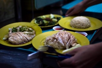 Several plates of food served on yellow plates. The central dish appears to be Hainanese chicken rice, featuring slices of poached chicken arranged on rice. Another plate includes leafy green vegetables. Small dishes of red sauce accompany the meal, and hands are seen setting down the plates.
