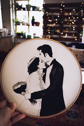 A hand holding an embroidery hoop featuring a black and white silhouette of a couple in a romantic embrace. The background reveals a cozy room with shelves adorned with various potted plants and softly glowing string lights, creating a warm and intimate atmosphere.