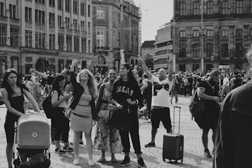 A bustling urban scene with many people gathered in a public square. A variety of individuals engage in different activities, such as taking selfies and pushing strollers. The background features historical and modern buildings. The crowd exudes a sense of liveliness, with some people seemingly celebrating or enjoying the moment.