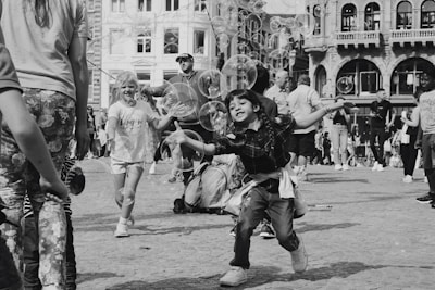 Children and adults are gathered in an outdoor urban area, enjoying a playful moment with floating bubbles. A child is prominently reaching out with excitement to catch the bubbles. The scene is lively, with people casually dressed and buildings in the background.