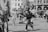 Children and adults are gathered in an outdoor urban area, enjoying a playful moment with floating bubbles. A child is prominently reaching out with excitement to catch the bubbles. The scene is lively, with people casually dressed and buildings in the background.
