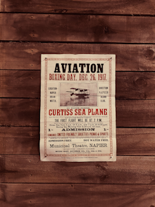 A vintage aviation poster from December 26, 1917, advertising a Curtiss Sea Plane event in Napier. The poster includes details such as the event's location, time, and admission information. It has a decorative border and is printed on aged paper, adding to its antique appearance. The poster is displayed on a wooden surface.