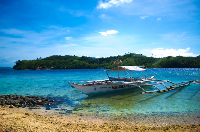 A vibrant outrigger boat gliding over crystal-clear waters surrounded by lush green Caramoan islands under a bright blue sky.