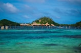 A tropical island with lush greenery and several thatched huts is surrounded by clear blue water. Wooden stilts can be seen in the water near the island. The sky is partly cloudy, adding a serene and peaceful backdrop.
