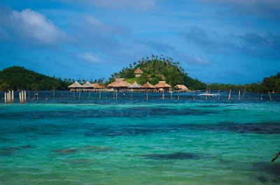 A tropical island with lush greenery and several thatched huts is surrounded by clear blue water. Wooden stilts can be seen in the water near the island. The sky is partly cloudy, adding a serene and peaceful backdrop.