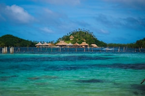 A tropical island with lush greenery and several thatched huts is surrounded by clear blue water. Wooden stilts can be seen in the water near the island. The sky is partly cloudy, adding a serene and peaceful backdrop.