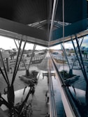 A modern architectural space with large metal beams and reflective glass panels. The central area features a walkway surrounded by greenery and seating areas. Several people are seen walking or sitting, and a series of trolley carts are neatly arranged. The scene extends into the distance, showing various structural elements and outdoor surroundings.