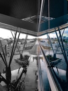 A modern architectural space with large metal beams and reflective glass panels. The central area features a walkway surrounded by greenery and seating areas. Several people are seen walking or sitting, and a series of trolley carts are neatly arranged. The scene extends into the distance, showing various structural elements and outdoor surroundings.