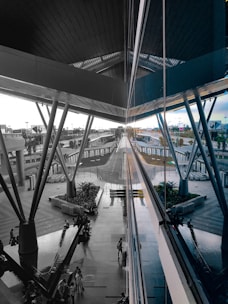 A modern architectural space with large metal beams and reflective glass panels. The central area features a walkway surrounded by greenery and seating areas. Several people are seen walking or sitting, and a series of trolley carts are neatly arranged. The scene extends into the distance, showing various structural elements and outdoor surroundings.