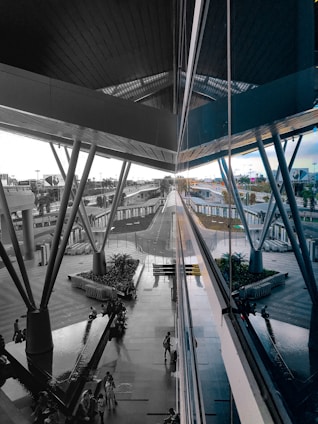 A modern architectural space with large metal beams and reflective glass panels. The central area features a walkway surrounded by greenery and seating areas. Several people are seen walking or sitting, and a series of trolley carts are neatly arranged. The scene extends into the distance, showing various structural elements and outdoor surroundings.