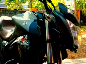 A close-up view of a motorcycle, focusing on the headlight, handlebars, and parts of the body. The motorcycle is parked outdoors with greenery and some blurred background elements visible. The brand logo is prominently displayed on the side.