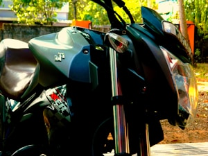 A close-up view of a motorcycle, focusing on the headlight, handlebars, and parts of the body. The motorcycle is parked outdoors with greenery and some blurred background elements visible. The brand logo is prominently displayed on the side.