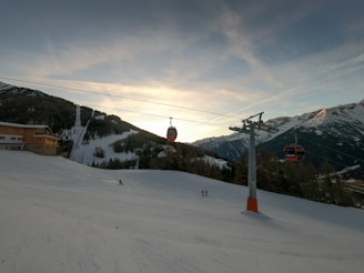 A scenic mountain landscape with snow-covered slopes and a sunset sky. Visible are ski lift cables with red gondolas suspended above the snowy terrain. A few skiers are dispersed across the slope, and a wooden lodge is positioned to the left surrounded by pine trees. Large mountain peaks rise in the background.