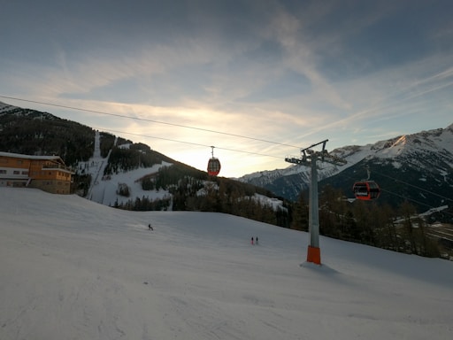 A scenic mountain landscape with snow-covered slopes and a sunset sky. Visible are ski lift cables with red gondolas suspended above the snowy terrain. A few skiers are dispersed across the slope, and a wooden lodge is positioned to the left surrounded by pine trees. Large mountain peaks rise in the background.