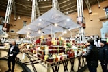 A bustling indoor market scene with several people interacting around a vendor stall displaying a wide variety of preserved goods, such as jars of pickles, sauces, and jams. The market is spacious, with a high ceiling and ample lighting. The structure is supported by metal beams, and there are decorative elements on the walls.