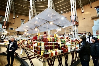 A bustling indoor market scene with several people interacting around a vendor stall displaying a wide variety of preserved goods, such as jars of pickles, sauces, and jams. The market is spacious, with a high ceiling and ample lighting. The structure is supported by metal beams, and there are decorative elements on the walls.