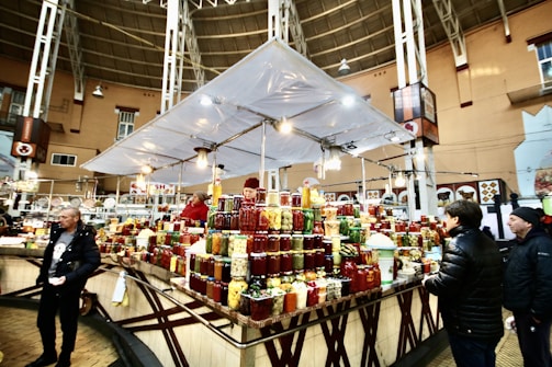 A bustling indoor market scene with several people interacting around a vendor stall displaying a wide variety of preserved goods, such as jars of pickles, sauces, and jams. The market is spacious, with a high ceiling and ample lighting. The structure is supported by metal beams, and there are decorative elements on the walls.